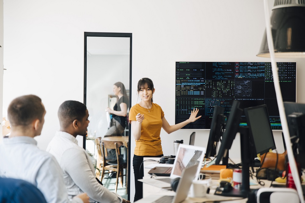 An IT worker is in a bright, modern office with two colleagues. She is standing at the front of a screen showing code and explaining something.