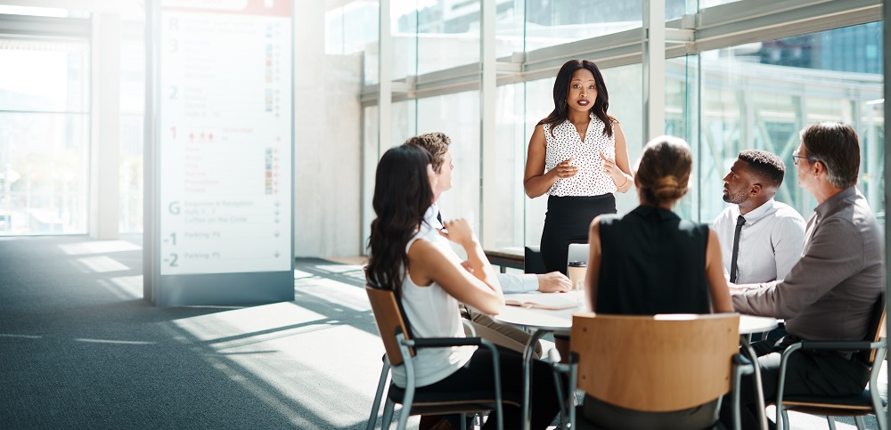 A diverse team sits at a round wooden table in a large, wide meeting room. One team member is giving a presentation.