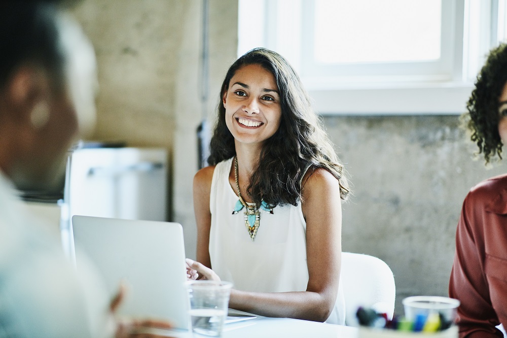 Woman sitting at her laptop with a bright smile. Her gaze is directed forward.